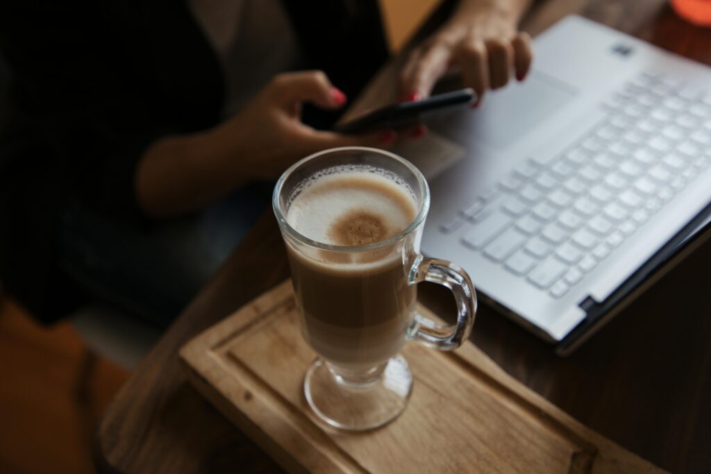 Person using a smartphone near a laptop with a latte on a wooden tray, creating a cozy work atmosphere.